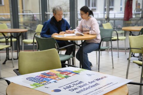 Eine ältere und eine jüngere Person sitzen an einem Tisch und lernen. Im Vordergrund liegt ein Plakat mit der Aufschrift "ABC-Treff. Die Welt der Worte entdecken!".