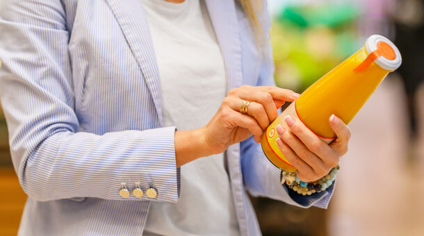 Eine Frau in hellblauem Jacket hält eine Flasche mit Saft in der Hand und führt beim lesen der Zutatenliste den Zeigefinger der anderen Hand mit. Das Gesicht ist nicht zu sehen.