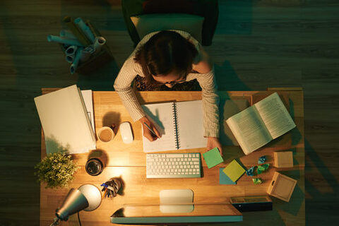 Schreibtisch von oben: eine Frau sitzt an einem computer uns schreibt in einen Block. Um sie herum lieben Bücher, Zettel, Blätter. Die Schreibtischlampe brennt, ringsherum ist es dunkel.